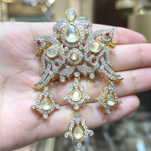 Può includere: Un ciondolo color oro raffigurante due figure di uccelli rivolte verso l'esterno, incastonato con numerose piccole pietre chiare e accentuato da due piccole pietre rosse. Il pezzo include tre castoni pendenti a forma di goccia.