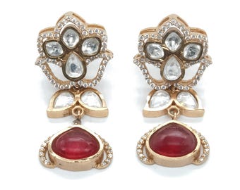 May include: A pair of gold-toned drop earrings featuring a floral-inspired top section encrusted with clear stones and a central teardrop-shaped red stone dangling from the base. The metal frame is detailed with small clear stones along the edges.