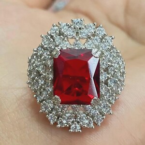 May include: A silver ring with a large, square, ruby-red gemstone at its centre, encircled by a halo of smaller, clear, round gemstones. The ring's design is detailed and striking, suitable for special events.