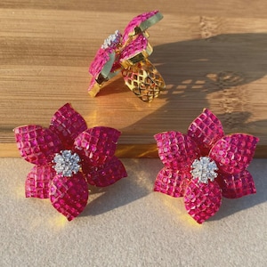 May include: A three-piece jewellery set with flower designs. The set includes a ring and two earrings. The flowers are a vibrant pink with a central cluster of white stones. The ring and earring bases are gold-coloured.