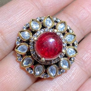 May include: A gold-toned ring with a large, round, red gemstone at the center. The ring is surrounded by smaller, clear gemstones and gold accents, creating a floral design. The ring is a statement piece.