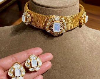 May include: A gold-toned mesh choker necklace featuring a central rectangular iridescent stone surrounded by smaller clear stones, with matching earrings. The jewelry set displays intricate metalwork and a polished finish throughout the design.