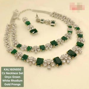May include: A Cz necklace set with onyx green stones and white rhodium accents. The necklace and matching earrings feature gold prongs and a floral design. The set includes a necklace and a pair of earrings.