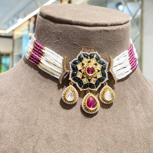 Puede incluir: Un collar gargantilla con un colgante floral. El colgante presenta una gema roja central, detalles en negro y dorado, y tres gemas en forma de lágrima. El collar tiene cuentas blancas y magenta.