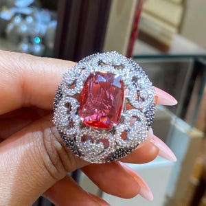 May include: A large, ornate ring featuring a rectangular, reddish-pink gemstone at its center. The gemstone is surrounded by a halo of small, sparkling white stones and a border of black and white stones. The ring is set on a silver band.