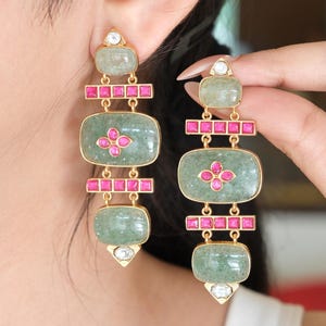 May include: A pair of gold-toned drop earrings featuring three rectangular, translucent light green stones and two horizontal bars of small, square, vibrant pink stones. A small, clear, triangular stone accents the top and bottom of each earring.