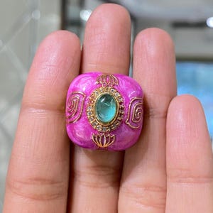 Puede incluir: Un anillo rosa vibrante con una cara cuadrada, con una piedra preciosa ovalada de color verde claro en el centro. La piedra preciosa está rodeada por un engaste dorado con pequeñas piedras transparentes. Detalles de filigrana dorada adornan los lados del anillo.