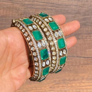Puede incluir: Dos brazaletes de oro con piedras verde esmeralda y transparentes. Los brazaletes están decorados con diseños intrincados y están engastados con una variedad de piedras.
