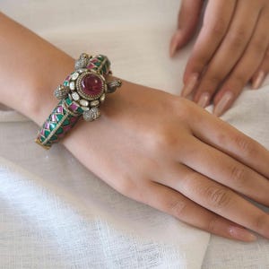 May include: A hinged metal bangle bracelet featuring a central turtle motif adorned with a large, round, deep pink cabochon stone, surrounded by small white stones. The band displays a geometric pattern of alternating green and pink enamel.
