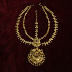 May include: A gold-toned, two-tiered headpiece with a large, ornate pendant featuring peacocks and purple gemstones. The headpiece is designed for traditional Indian weddings.