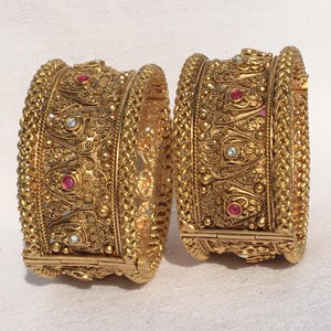 Puede incluir: Dos brazaletes dorados ornamentados. Cada brazalete presenta intrincados patrones en relieve, incluyendo diseños florales y geométricos. Pequeñas piedras preciosas rojas y claras están engastadas en el diseño. Los brazaletes tienen un cierre de bisagra.