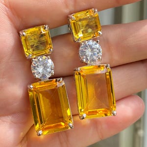 Puede incluir: Un par de pendientes plateados con un diseño de tres piedras. Cada pendiente presenta una gema amarilla cuadrada en la parte superior, una gema clara redonda en el medio y una gema amarilla rectangular en la parte inferior. Las gemas están engastadas en metal plateado.
