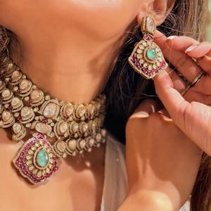 Peut inclure: Gros plan sur un ensemble de bijoux doré luxueux comprenant un collier et des boucles d'oreilles. Le collier et les boucles d'oreilles présentent un motif floral avec des pierres claires et de couleur rubis, et une pierre verte centrale. Les boucles d'oreilles sont tenues.