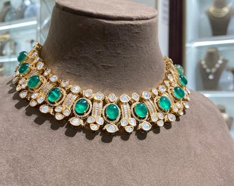 May include: A gold-toned necklace with large, oval emeralds and clear, round and baguette-cut gemstones. The necklace is displayed on a brown mannequin head. The necklace has a floral design.