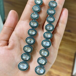 Pu&ograve; includere: Un paio di orecchini pendenti lunghi. Ogni orecchino presenta una serie di pietre preziose chiare di forma ovale, circondate da piccole pietre color teal. Gli orecchini sono incastonati in un metallo argentato.