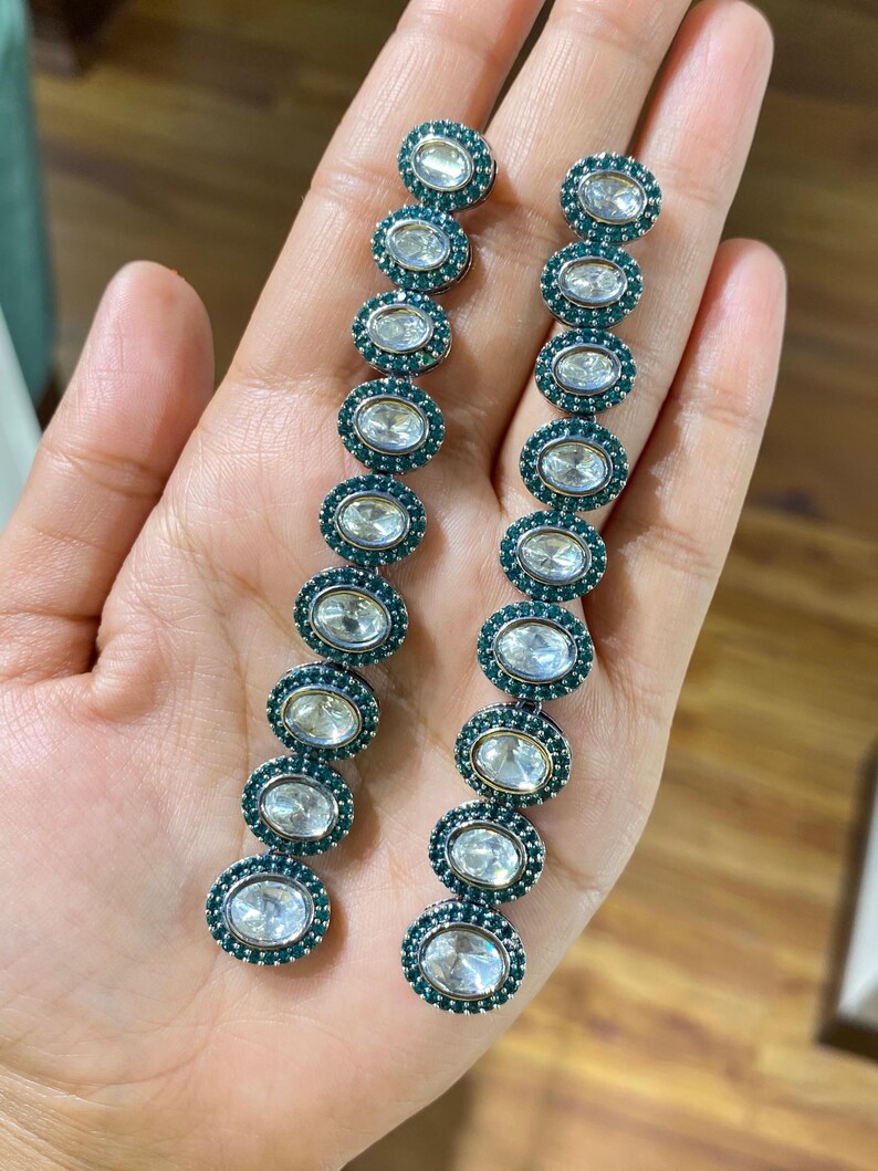 Pu&ograve; includere: Un paio di orecchini pendenti lunghi. Ogni orecchino presenta una serie di pietre preziose chiare di forma ovale, circondate da piccole pietre color teal. Gli orecchini sono incastonati in un metallo argentato.