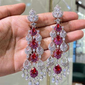 May include: Elegant chandelier earrings with a silver-tone metal setting. The earrings feature a cascade of clear and ruby-red teardrop-shaped stones, accented with smaller clear stones. The top of the earrings has a leaf-like design.