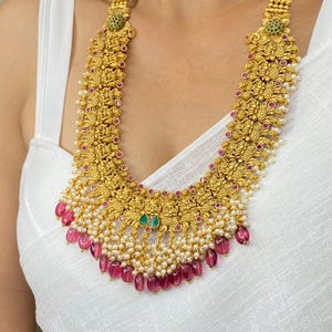 May include: A gold-toned necklace with intricate floral and figure details. The necklace features small red and green gemstones, white pearls, and dangling pink beads. The necklace is worn against a white background.
