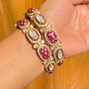 May include: Gold bangles featuring alternating oval and round gemstones. The oval stones are clear, and the round stones are a deep red. The bangles are embellished with small, clear accent stones, creating a luxurious appearance.