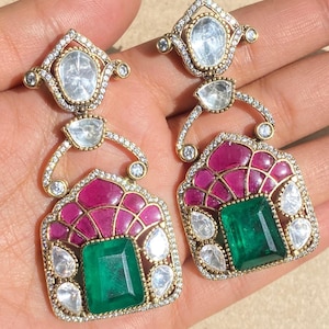 May include: A pair of ornate earrings featuring a gold-tone setting. Each earring has a large, rectangular emerald-green stone at the center, surrounded by smaller clear stones and a pink, fan-shaped design. The earrings are embellished with additional clear stones.