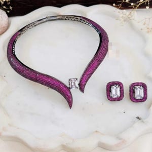 Könnte beinhalten: Ein Set aus magentafarbenem Halsband und Ohrringen. Die Halskette ist im Kragenstil gehalten und mit kleinen magentafarbenen Steinen besetzt. Die Ohrringe sind quadratisch mit einem großen, klaren Stein in der Mitte, umgeben von magentafarbenen Steinen.