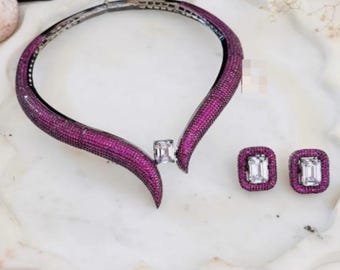 May include: A magenta-colored necklace and matching earrings set. The necklace is a collar style, encrusted with small magenta stones. The earrings are square-shaped with a large, clear stone in the center, surrounded by magenta stones.