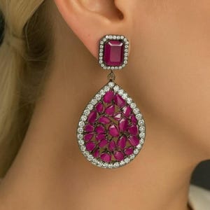 Puede incluir: Pendientes colgantes con una gema rectangular de color rojo intenso rodeada de pequeñas piedras transparentes. La parte inferior en forma de lágrima está llena de gemas rojas oscuras más pequeñas, también enmarcadas por piedras transparentes. Montura de metal oscuro.