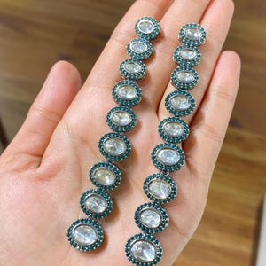 Pu&ograve; includere: Un paio di orecchini pendenti lunghi. Ogni orecchino presenta una serie di pietre ovali e trasparenti circondate da un'incastonatura color verde acqua. Gli orecchini sono esposti su uno sfondo neutro, evidenziando il loro design e colore.