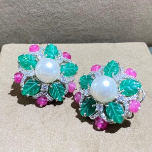 May include: A pair of floral-shaped earrings. Each earring features a large white pearl center, surrounded by green leaf-shaped accents, pink gemstone clusters, and small clear crystals. The earrings have a silver-colored metal setting.