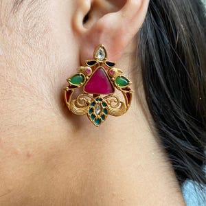 Puede incluir: Pendiente de botón dorado con una piedra magenta triangular en el centro. El pendiente presenta detalles en verde, blanco y rojo, con un diseño detallado y ornamentado. El pendiente se lleva en una oreja.
