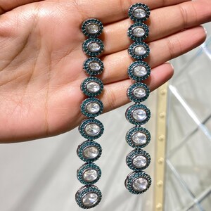 Pu&ograve; includere: Un paio di orecchini pendenti lunghi. Ogni orecchino presenta una serie di pietre chiare di forma ovale circondate da un'incastonatura verde scuro. Gli orecchini sono tenuti in mano, mostrando la loro lunghezza e il loro design.