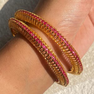 May include: Two gold-colored bangles with a repeating pattern of small, round, pink gemstones. The bangles have an openwork design with a textured surface. The gemstones are set in a row along the center of the bangles.
