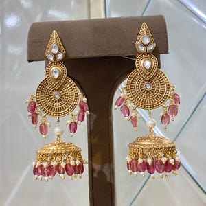 Könnte beinhalten: Vergoldete Ohrringe mit aufwendigen Details. Jeder Ohrring hat eine tropfenförmige Spitze mit klaren Steinen, ein kreisförmiges Design mit einem zentralen Stein und hängenden rosa Edelsteinen und Perlen. Der untere Teil hat eine Glockenform.