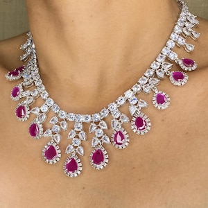 Puede incluir: Un collar deslumbrante con un engaste de metal plateado adornado con numerosas piedras claras y brillantes. El collar est&aacute; acentuado con vibrantes piedras preciosas de color rojo rub&iacute; en forma de l&aacute;grima, creando un dise&ntilde;o lujoso y llamativo.