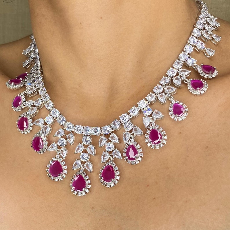 Puede incluir: Un collar deslumbrante con un engaste de metal plateado adornado con numerosas piedras claras y brillantes. El collar est&aacute; acentuado con vibrantes piedras preciosas de color rojo rub&iacute; en forma de l&aacute;grima, creando un dise&ntilde;o lujoso y llamativo.