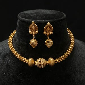 May include: A gold choker necklace with a large round bead in the center and two smaller round beads on either side. The necklace is made of small, round beads. The necklace comes with a pair of matching earrings with a teardrop shape and a round bead at the bottom.