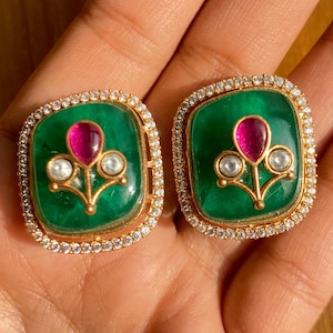 Pode incluir: Um par de brincos quadrados. Cada brinco apresenta uma vibrante pedra central verde, uma gema rosa central e duas pérolas brancas, tudo em metal dourado. Os brincos são emoldurados por uma borda de pequenas pedras transparentes.