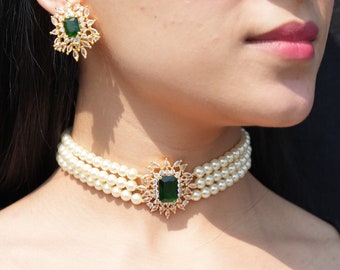May include: A three-strand pearl choker necklace with a gold and green gemstone pendant. The pendant features a square-shaped green gemstone surrounded by smaller clear gemstones.