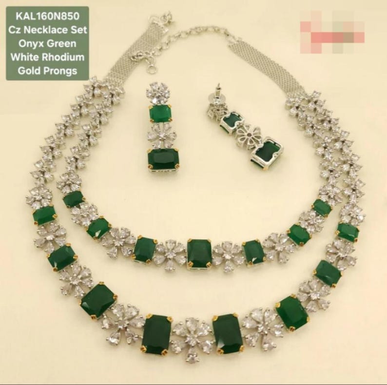 May include: A Cz necklace set with onyx green stones and white rhodium plating. The set includes a necklace and matching earrings. The necklace features a double-strand design with emerald-cut green stones and floral accents. The earrings have a similar design.