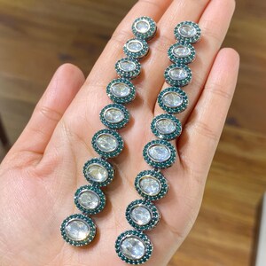 Pu&ograve; includere: Un paio di orecchini pendenti lunghi. Ogni orecchino presenta una serie di pietre chiare di forma ovale, circondate da un anello di piccole pietre color verde acqua. Gli orecchini sono tenuti da una mano, mostrando il loro design e la loro lucentezza.