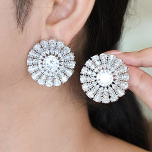 May include: A pair of large, circular stud earrings featuring a central round-cut clear stone surrounded by two concentric rings of smaller, pear-shaped clear stones set in a silver-toned metal frame, creating a radiant, floral-like pattern.