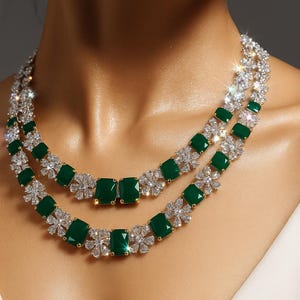 May include: A two-strand necklace featuring emerald-cut green gemstones and sparkling clear crystals. The necklace has a gold-colored setting and a floral design between the green stones. The jewelry is worn around the neck.