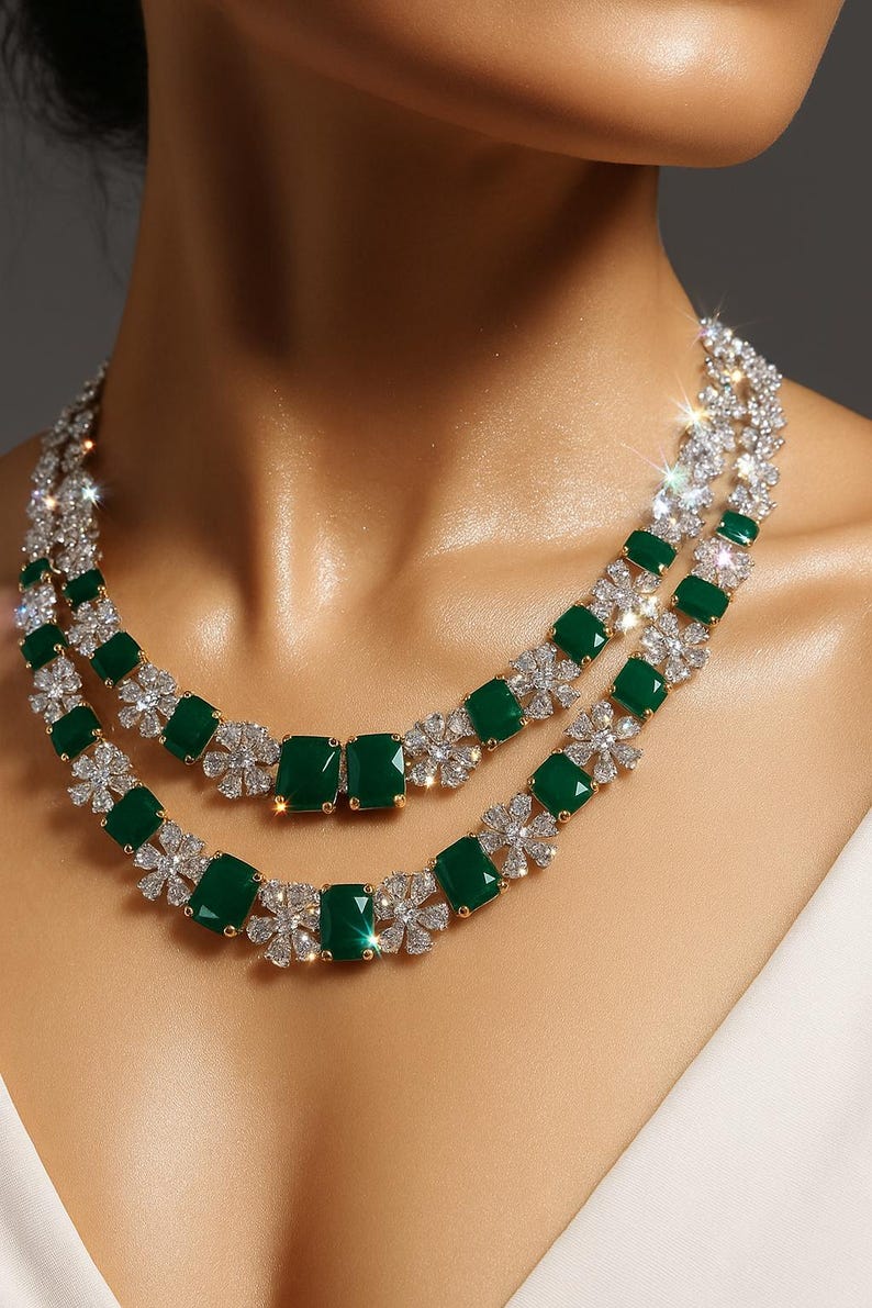 May include: A two-strand necklace featuring emerald-cut green gemstones and sparkling clear crystals. The necklace has a gold-colored setting and a floral design between the green stones. The jewelry is worn around the neck.