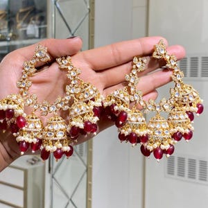 Könnte beinhalten: Ein Paar goldfarbene Chandelier-Ohrringe mit aufwendigen Blumen- und Blattmustern. Die Ohrringe haben klare Kristalle und hängende rote Perlen mit kleinen weißen Perlen. Die Ohrringe werden von einer Hand gehalten.
