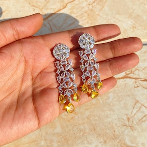 May include: A pair of silver dangle earrings with clear and yellow cubic zirconia stones. The earrings feature a flower design with multiple tiers of stones.