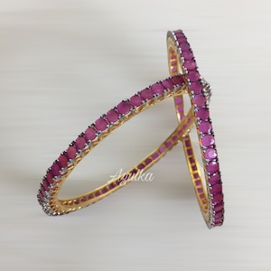 May include: Two gold bangles, each adorned with a line of magenta gemstones set in silver-coloured metal. The bangles are displayed on a light surface. The brand name "Agulka" is visible.