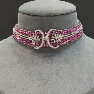 May include: A magenta beaded choker necklace with a silver clasp and floral detailing. The necklace is displayed on a grey mannequin head, highlighting the intricate design and colour of the jewellery.