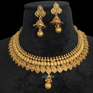 May include: A gold-colored necklace and earring set. The necklace features a detailed design with leaf-shaped pendants and small beads. The earrings have a bell-like shape with a dangling sphere. The set is displayed on a black surface.