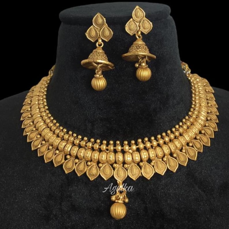 May include: A gold-colored necklace and earring set. The necklace features a detailed design with leaf-shaped pendants and small beads. The earrings have a bell-like shape with a dangling sphere. The set is displayed on a black surface.