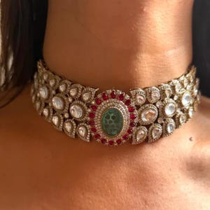 May include: A silver-toned choker necklace with an oval green center stone surrounded by small red stones. The necklace features clear, round and teardrop-shaped stones.